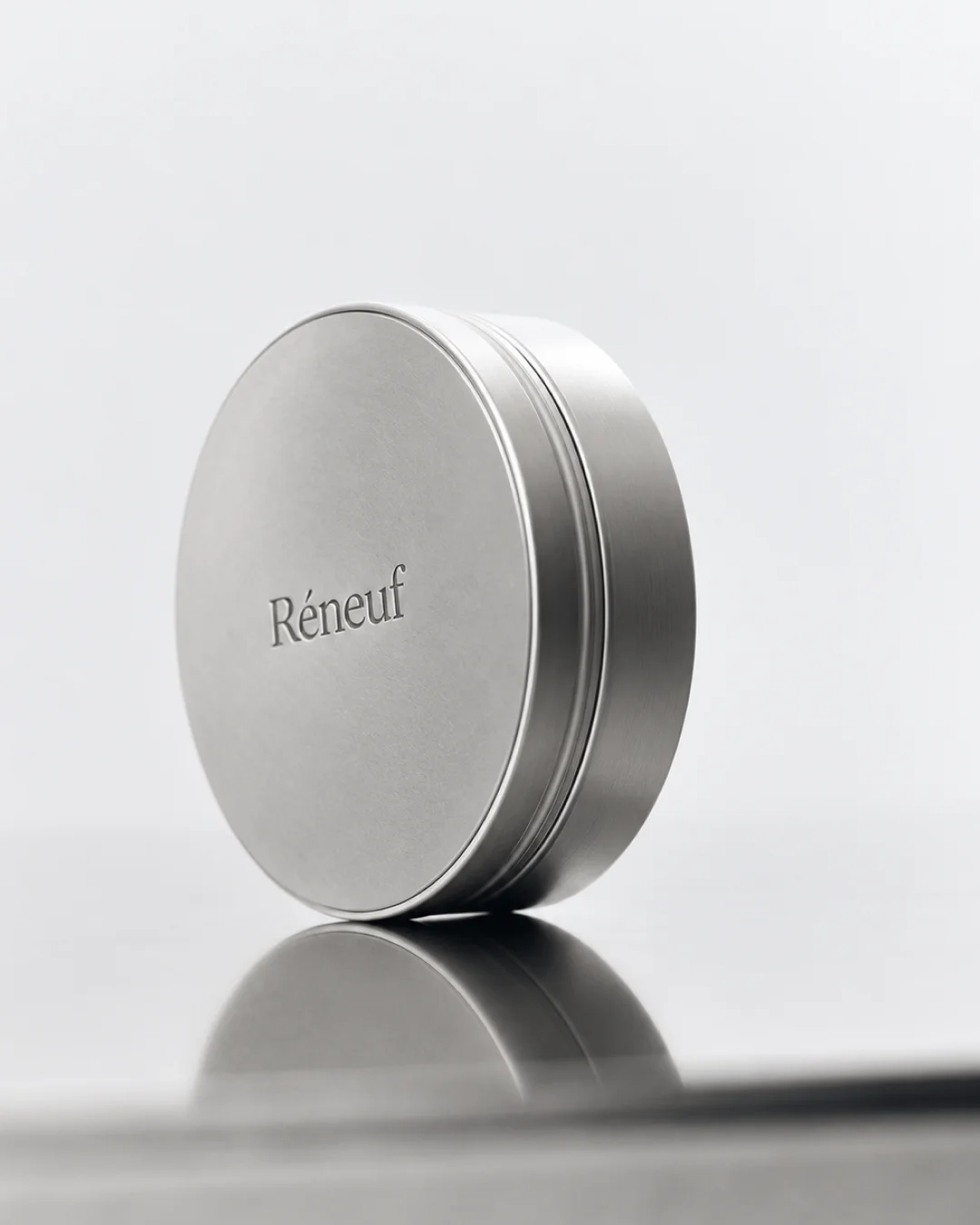Round silver Réneuf skincare tin photographed upright on a reflective surface.