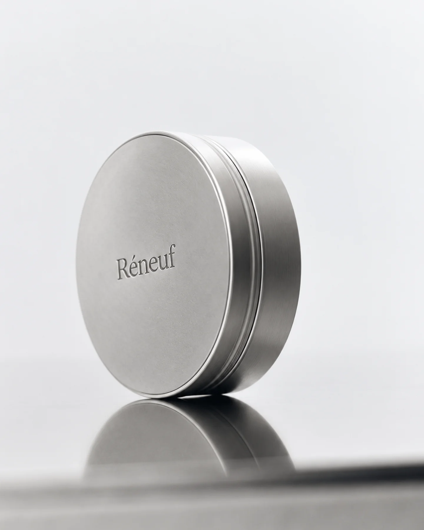 Round silver Réneuf skincare tin photographed upright on a reflective surface.
